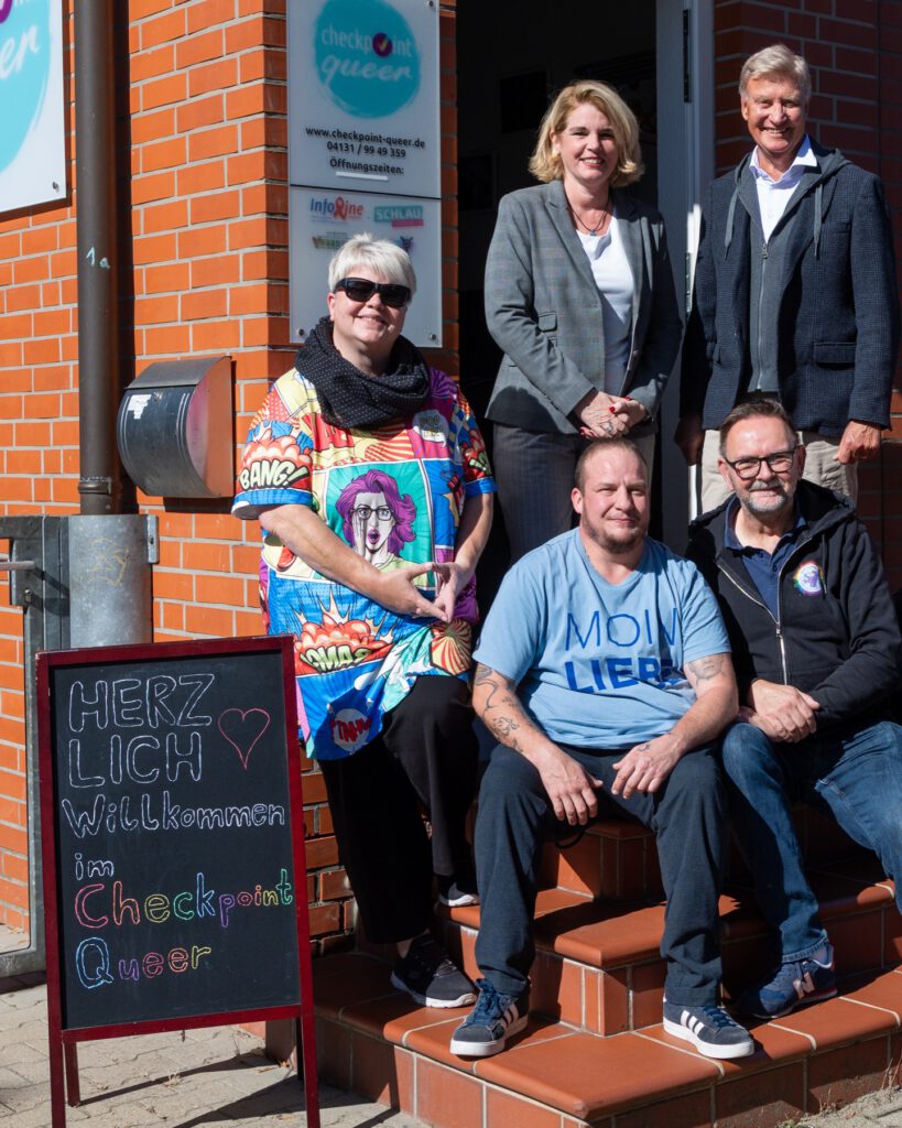 Claudia Kalisch und Ole von Beust mit dem Team des Checkpoint Queer vor dem Eingang, davor eine Tafel: Herzlich willkommen im Checkpoint Queer