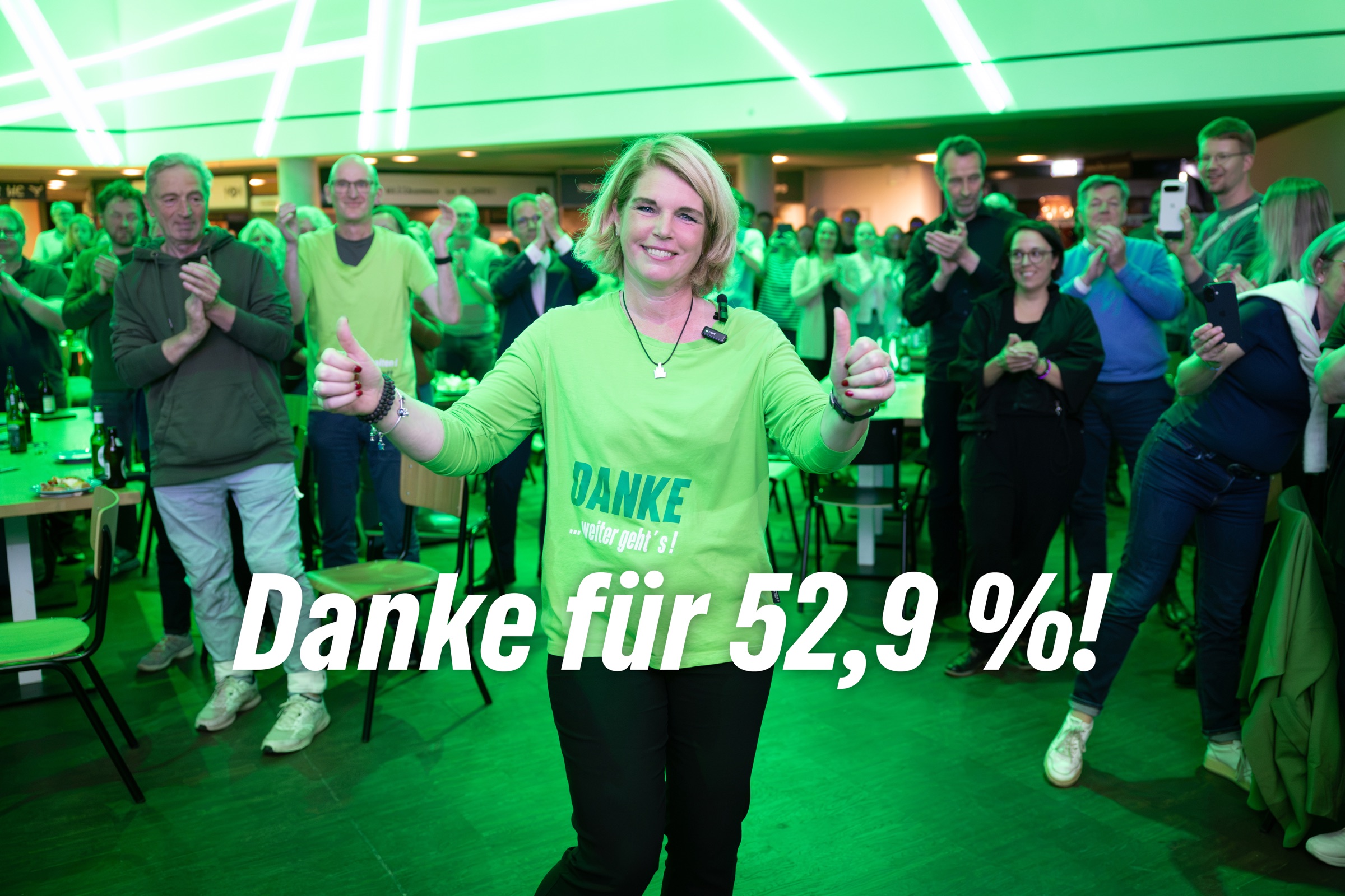 Claudia Kalisch reckt am Wahlabend beide Daumen hoch, hinter ihr applaudieren Unterstützer:innen – Danke für 52,9 %!